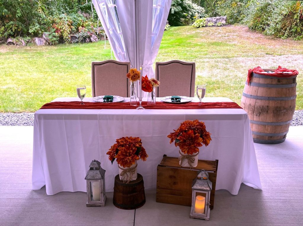 Sweetheart Table with floral arrangements, and lanterns under a white tent, set up for a reception at the 228 in Sterling, MA. Sunlight filters through the windowed tent walls.