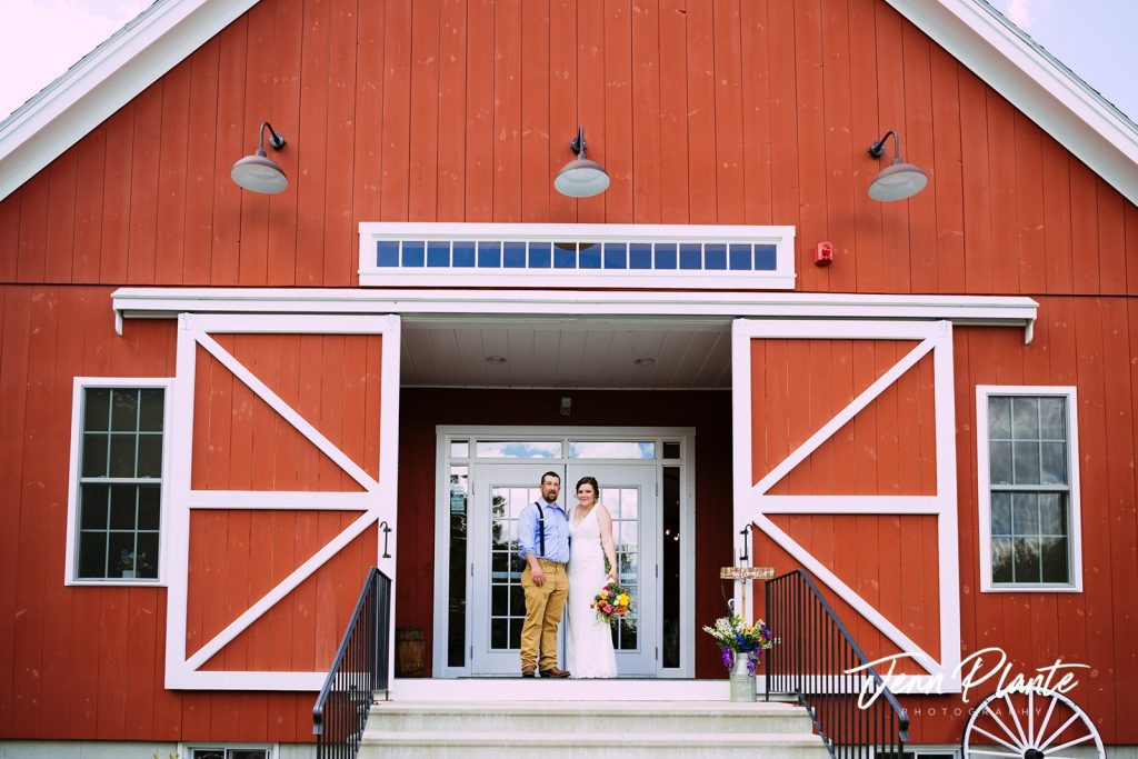 Couple standing on steps in front of a red barn with white doors, holding hands and smiling at the 228 in Sterling, MA